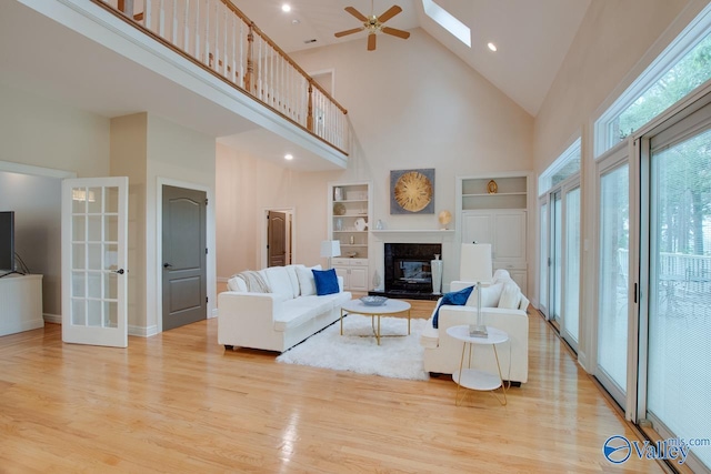 living room featuring high vaulted ceiling, built in shelves, a skylight, light wood-type flooring, and a glass covered fireplace