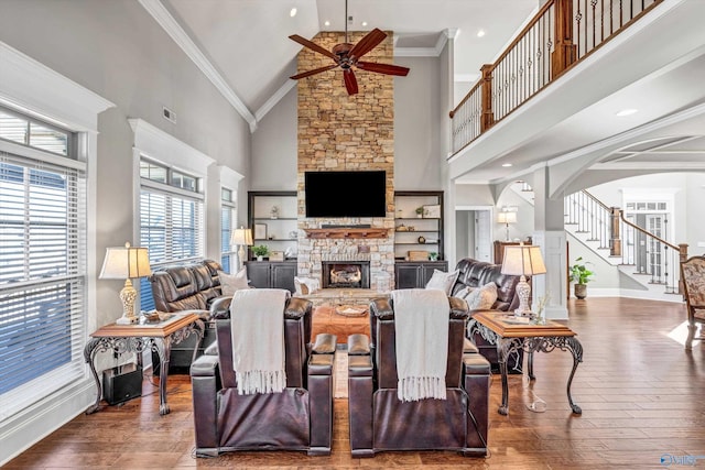 living area featuring arched walkways, a towering ceiling, wood-type flooring, ornamental molding, and a stone fireplace