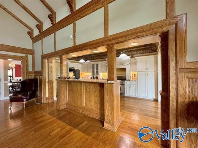 kitchen featuring a peninsula, a high ceiling, white cabinets, light wood finished floors, and decorative columns