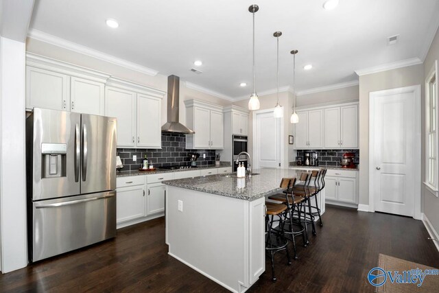 kitchen featuring light stone counters, appliances with stainless steel finishes, white cabinetry, an island with sink, and wall chimney exhaust hood