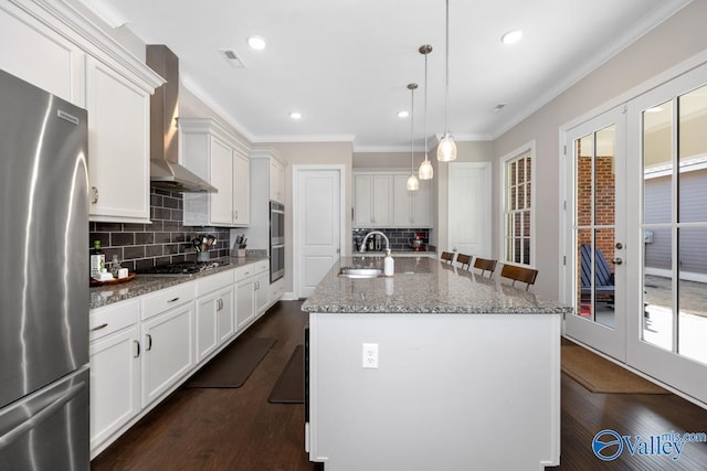 kitchen with decorative light fixtures, a center island with sink, stainless steel appliances, white cabinetry, and wall chimney exhaust hood