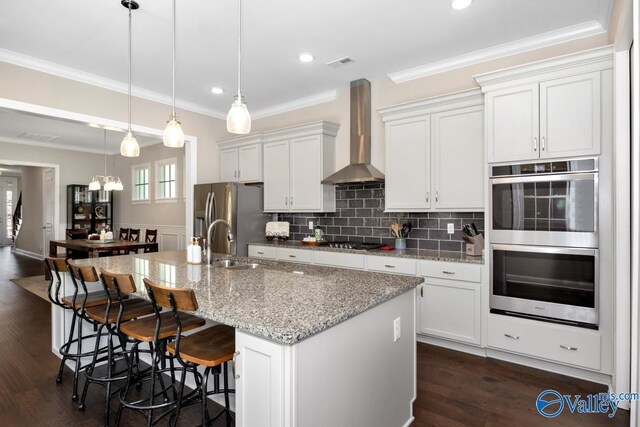 kitchen with decorative light fixtures, appliances with stainless steel finishes, white cabinets, wall chimney range hood, and an island with sink