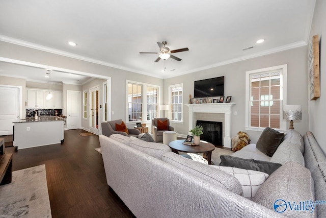 living area featuring ceiling fan, visible vents, ornamental molding, dark wood-style floors, and a glass covered fireplace