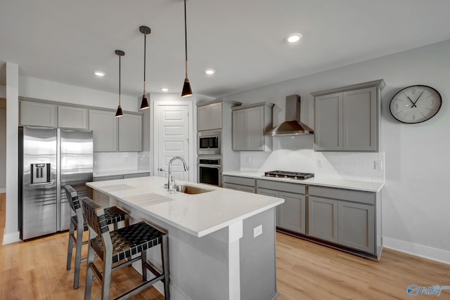 kitchen featuring wall chimney exhaust hood, a kitchen island with sink, stainless steel appliances, gray cabinetry, and a sink