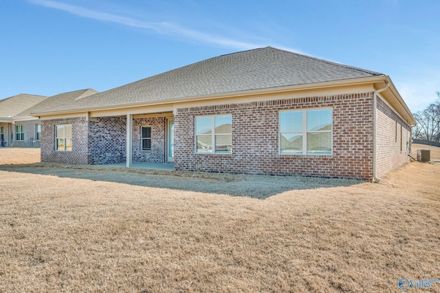 back of property featuring brick siding, a lawn, central AC, and roof with shingles