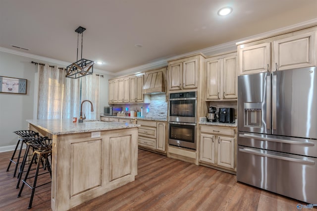 kitchen featuring premium range hood, appliances with stainless steel finishes, ornamental molding, a center island with sink, and decorative light fixtures