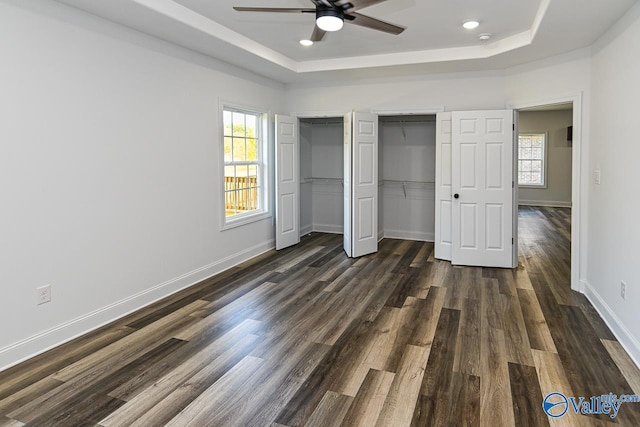 unfurnished bedroom featuring a tray ceiling, multiple windows, ceiling fan, and dark wood-type flooring