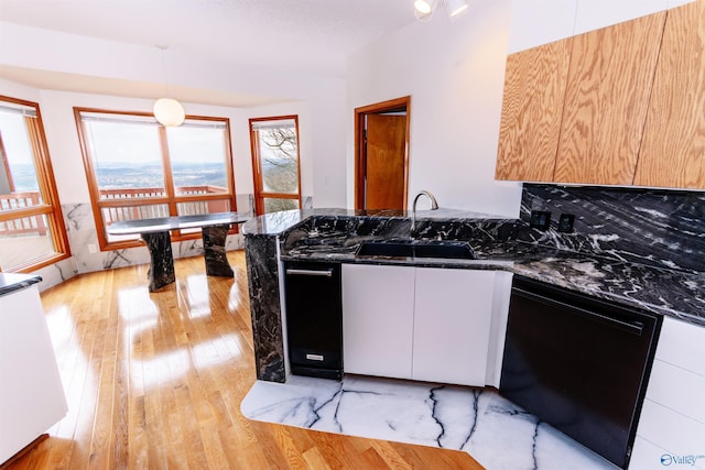 kitchen with black dishwasher, a sink, dark stone countertops, light wood-type flooring, and a peninsula