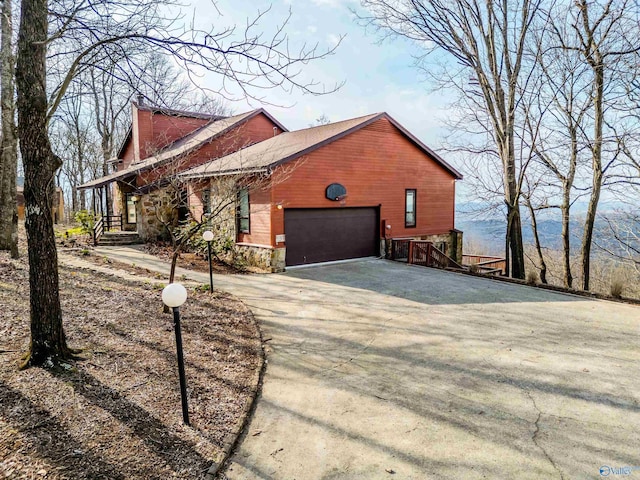 view of side of property featuring driveway, stone siding, an attached garage, and a chimney