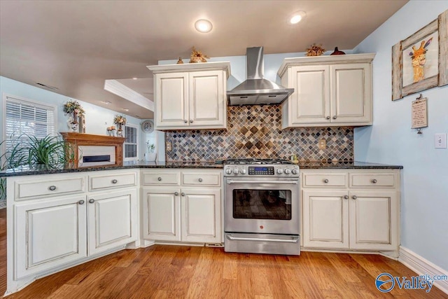 kitchen with light hardwood / wood-style floors, backsplash, dark stone countertops, stainless steel gas stove, and wall chimney range hood