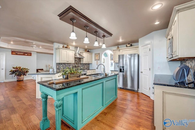 kitchen with a center island with sink, appliances with stainless steel finishes, decorative light fixtures, wall chimney exhaust hood, and sink