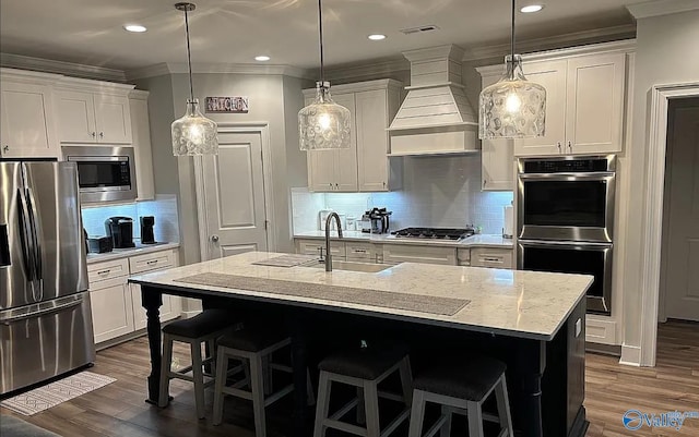 kitchen featuring white cabinetry, stainless steel appliances, an island with sink, pendant lighting, and custom range hood