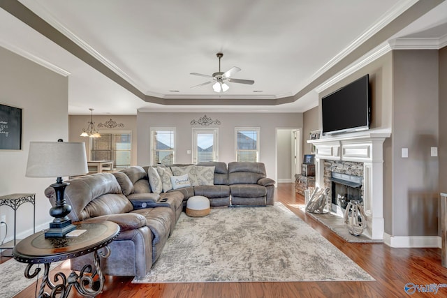 living area featuring crown molding, dark wood finished floors, a raised ceiling, a stone fireplace, and baseboards