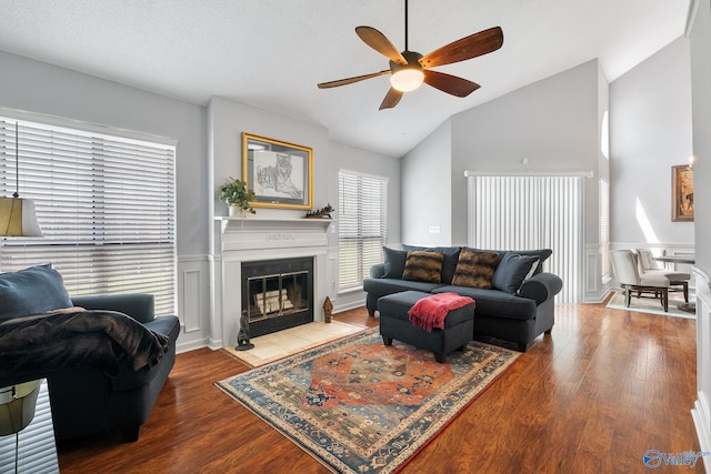 living area with lofted ceiling, ceiling fan, a fireplace with flush hearth, wood finished floors, and wainscoting