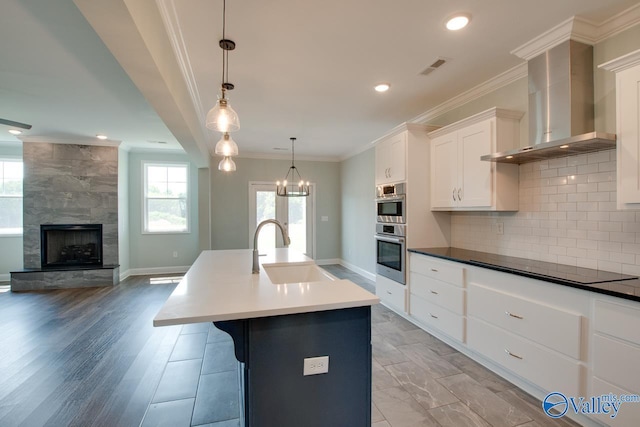 kitchen with sink, wall chimney range hood, an island with sink, a fireplace, and light wood-type flooring