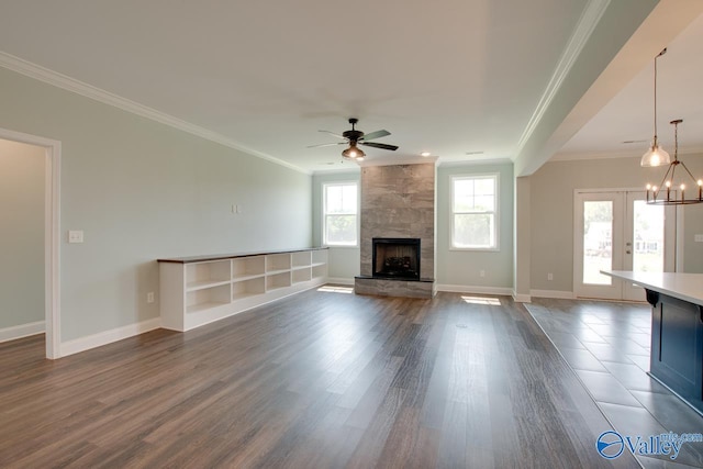 unfurnished living room featuring french doors, ceiling fan with notable chandelier, a stone fireplace, crown molding, and dark hardwood / wood-style floors