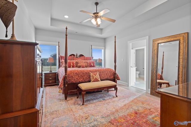 bedroom with a tray ceiling, ensuite bath, light wood-style floors, baseboards, and ceiling fan