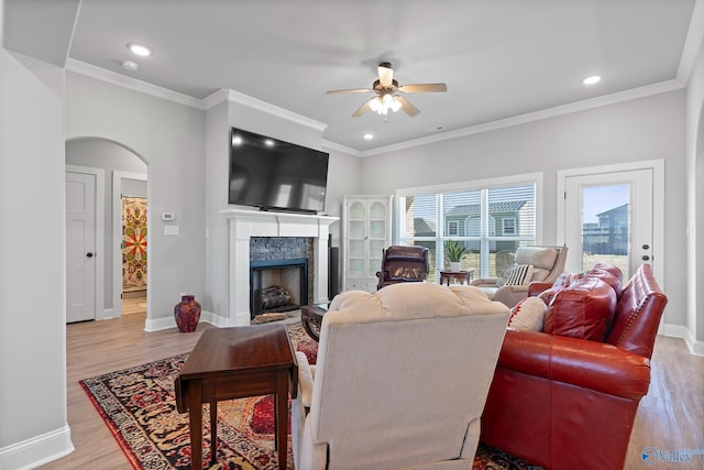 living area featuring ornamental molding, arched walkways, light wood-style floors, a stone fireplace, and baseboards