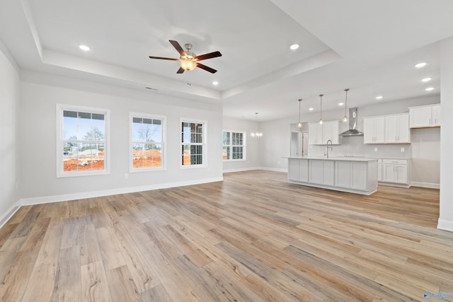 unfurnished living room with light wood finished floors, a tray ceiling, recessed lighting, and ceiling fan with notable chandelier