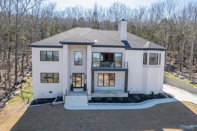 view of front of house with roof with shingles, a chimney, crawl space, a balcony, and a wooded view