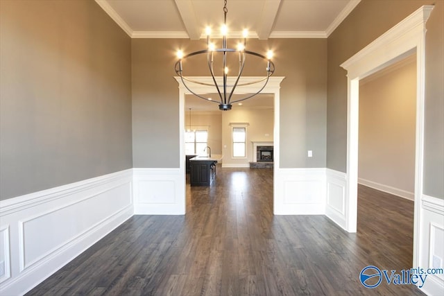 unfurnished dining area with crown molding, beam ceiling, a notable chandelier, dark hardwood / wood-style flooring, and a stone fireplace