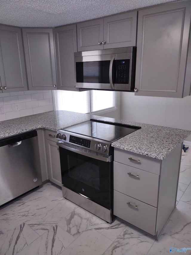 kitchen with appliances with stainless steel finishes, a textured ceiling, gray cabinetry, light stone countertops, and marble finish floor