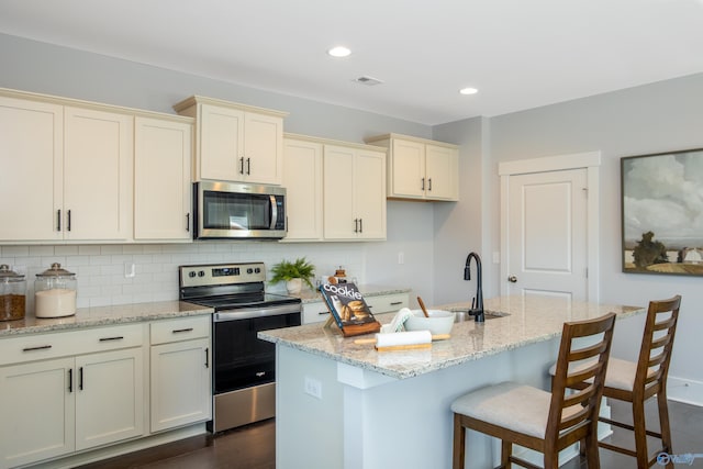 kitchen with light stone counters, a center island with sink, decorative backsplash, appliances with stainless steel finishes, and a sink