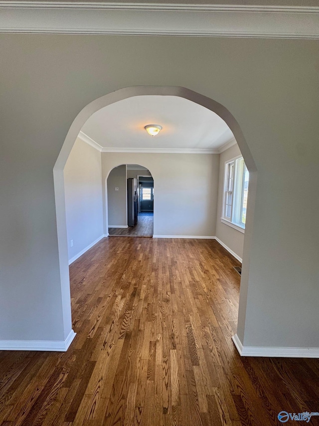 empty room featuring arched walkways, crown molding, baseboards, and wood finished floors