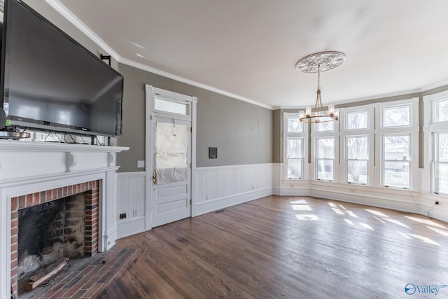 unfurnished living room with a notable chandelier, ornamental molding, wood finished floors, wainscoting, and a brick fireplace