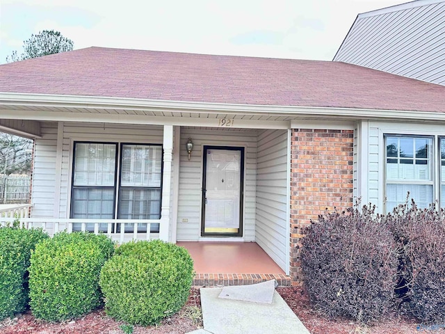 doorway to property featuring covered porch, brick siding, and roof with shingles