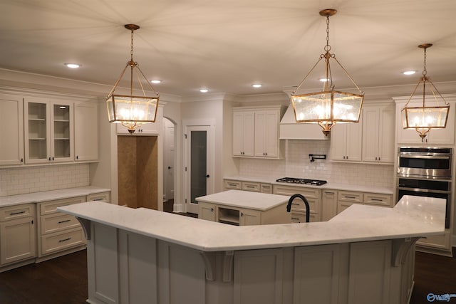 kitchen with crown molding, hanging light fixtures, stainless steel double oven, decorative backsplash, and a large island with sink