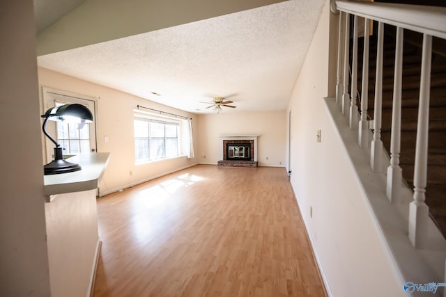 unfurnished living room with light wood finished floors, a ceiling fan, stairs, a textured ceiling, and a brick fireplace