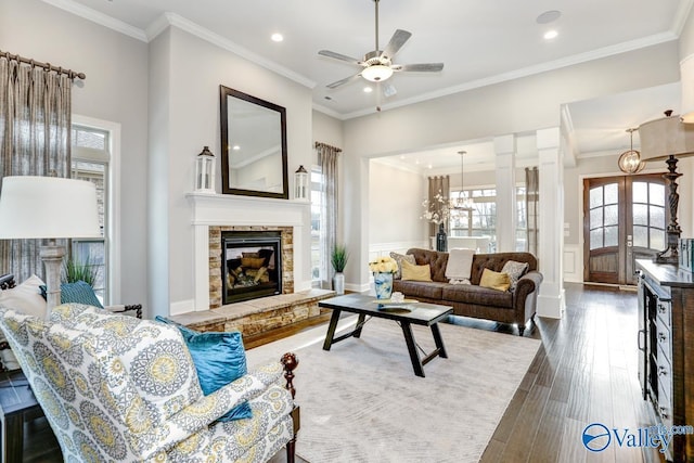 living room with dark wood-type flooring, french doors, a stone fireplace, ornamental molding, and ceiling fan with notable chandelier