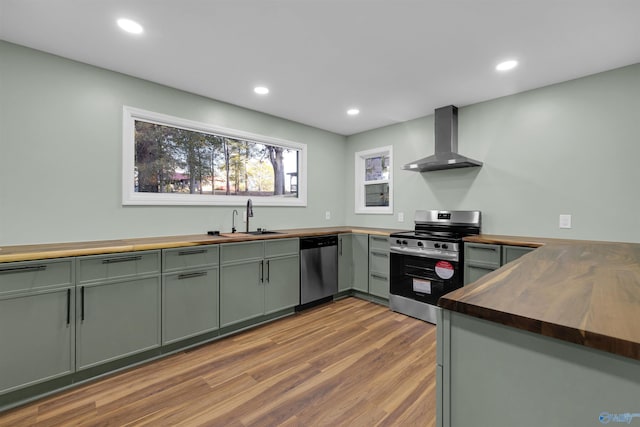 kitchen with wall chimney range hood, wood-type flooring, wood counters, sink, and appliances with stainless steel finishes