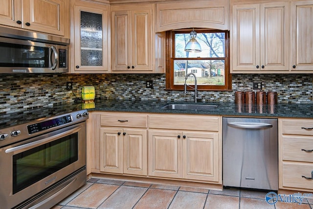 kitchen with stainless steel appliances, backsplash, glass insert cabinets, a sink, and dark stone countertops