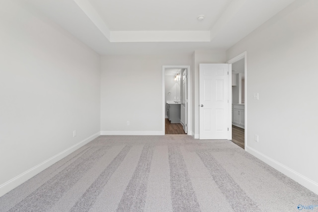 unfurnished bedroom with carpet floors, a tray ceiling, baseboards, and ensuite bathroom