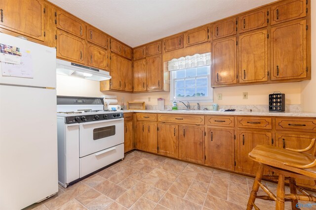 kitchen featuring light countertops, under cabinet range hood, brown cabinets, and white appliances