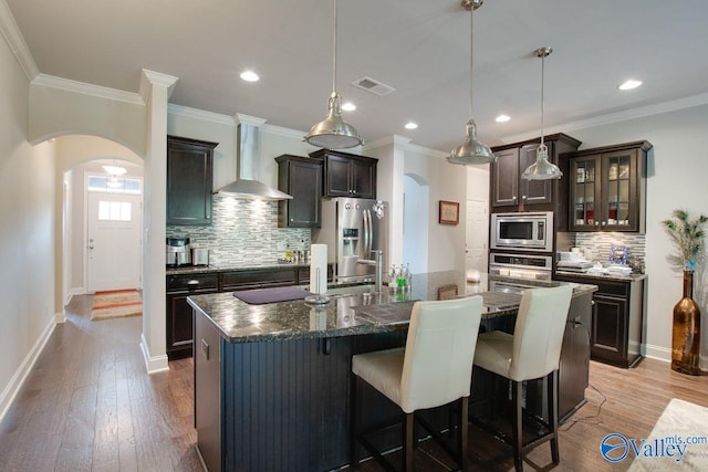 kitchen featuring wall chimney range hood, an island with sink, appliances with stainless steel finishes, light hardwood / wood-style flooring, and decorative light fixtures