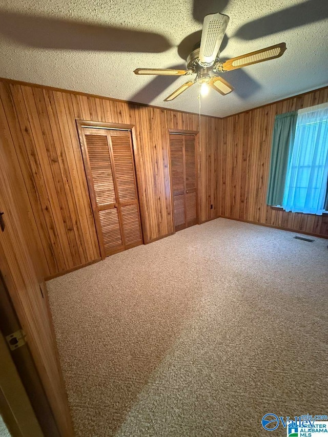 unfurnished bedroom featuring wooden walls, carpet, ceiling fan, multiple closets, and a textured ceiling