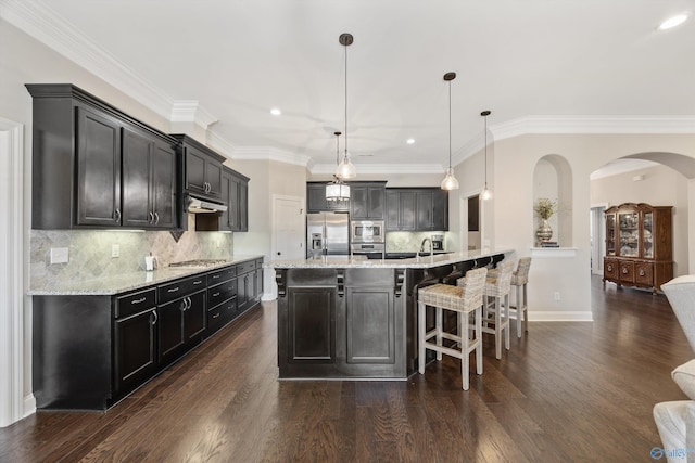 kitchen featuring arched walkways, hanging light fixtures, appliances with stainless steel finishes, a large island with sink, and under cabinet range hood