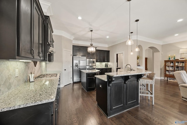 kitchen featuring stainless steel appliances, arched walkways, a kitchen island with sink, and decorative light fixtures