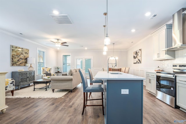 kitchen with wall chimney exhaust hood, sink, decorative light fixtures, electric range, and a kitchen island with sink