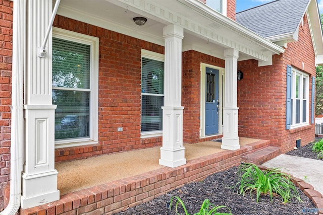 view of exterior entry featuring a porch, a shingled roof, and brick siding