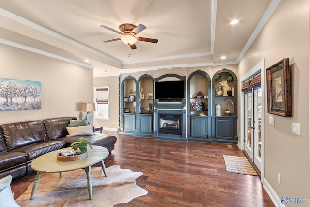 living area featuring baseboards, a raised ceiling, a glass covered fireplace, dark wood-style floors, and crown molding