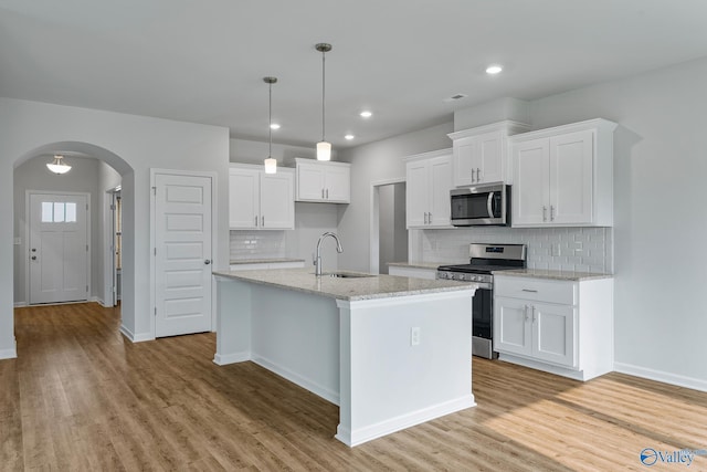 kitchen featuring arched walkways, a kitchen island with sink, stainless steel appliances, white cabinetry, and a sink