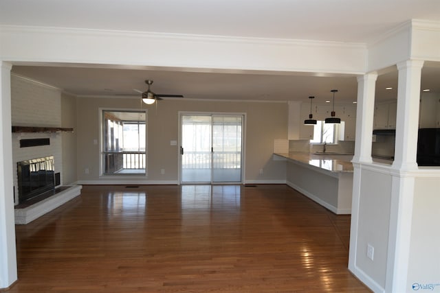 unfurnished living room featuring ceiling fan, decorative columns, ornamental molding, dark hardwood / wood-style flooring, and a brick fireplace