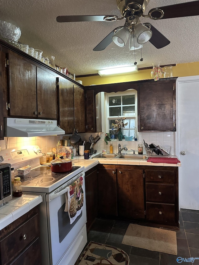 kitchen with white electric range, dark brown cabinetry, under cabinet range hood, tile countertops, and a sink