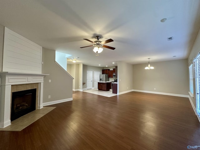 unfurnished living room with ceiling fan with notable chandelier, dark wood-style flooring, visible vents, baseboards, and a tiled fireplace