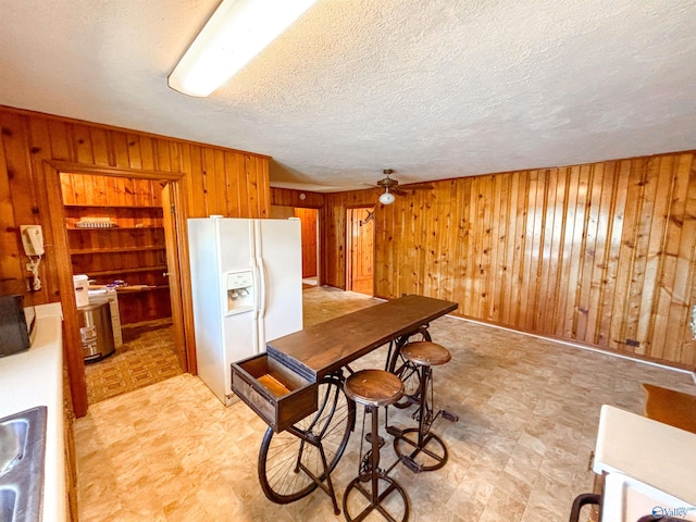 kitchen featuring a textured ceiling, wood walls, white refrigerator with ice dispenser, and ceiling fan