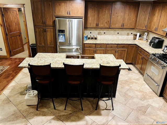 kitchen with brown cabinetry, light stone countertops, a breakfast bar, decorative backsplash, and appliances with stainless steel finishes